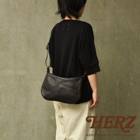 一位模特身著黑色上衣和米色褲子，斜挎一個黑色半月形單肩包。包袋由皮革製成，表面呈現自然的紋理，有金屬搭扣和肩帶調節扣。