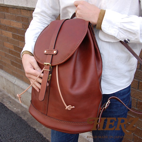 職人手作皮革單扣束口後背包，採用鞣製牛皮材質，呈現自然的棕色調。包款設計有單一束口與翻蓋式開口，並配有可調節的皮革肩帶和金屬扣環。