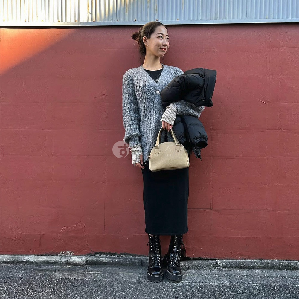 一位女性手持淺金色半月形迷你手提包，包身呈現皮革紋理，搭配灰色針織外套、黑色長裙和黑色厚底靴。