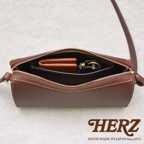 俯視視角拍攝一款日本HERZ品牌深褐色皮革單肩包，包口處敞開呈現內部結構，配有拉鍊開合與肩背帶，包內置有小型錢包及鑰匙扣等物品，圖片右下角印有HERZ品牌標誌與手工製造年份字樣。