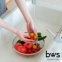 一個不鏽鋼圓形網狀瀝水籃，籃中裝有番茄和黃瓜。一雙手正在水龍頭下沖洗小番茄，水流從龍頭流出。