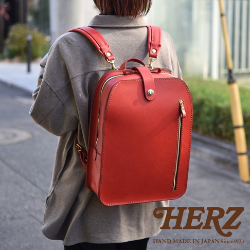 紅色皮革後背包，附有金屬拉鍊和扣環，設計簡約，可肩背或手提。