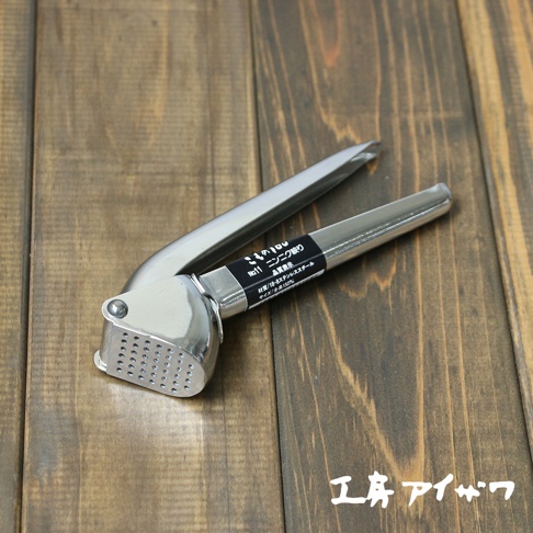 不鏽鋼壓蒜器，帶有銀色金屬光澤，壓蒜部位有圓形孔洞，手柄為流線型設計，置於木紋背景上。