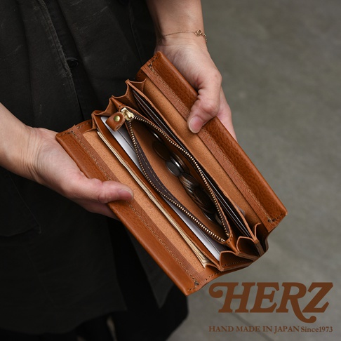 一人手持一款日本HERZ職人手作皮革長夾，錢包內層設計有拉鍊零錢袋並放入硬幣，同時可收納紙鈔與卡片，展現質感極佳的焦糖色皮革紋理與細緻車縫工藝，右下角印有HERZ品牌字樣。