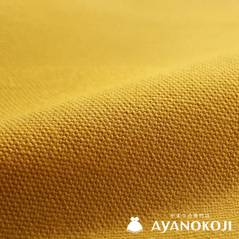一塊黃色的帆布織物，呈現出粗糙的紋理，並帶有褶皺，右下角有「AYANOKOJI」的品牌標誌。