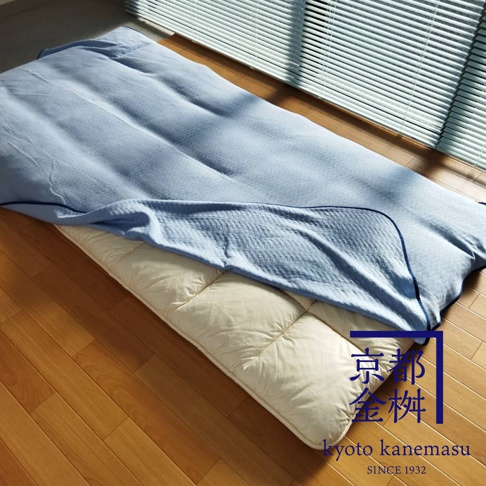 一張淺藍色天絲棉絎縫兩用單人毯，鋪在米白色鋪棉床墊上，床墊放在木地板上，背景是百葉窗。