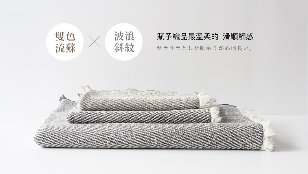 堆疊的長毛巾，呈現雙色流蘇和波浪斜紋織紋，材質看來柔軟，有深淺灰白色調。