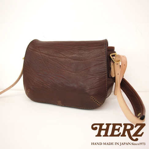 深棕色皮革郵差包，帶有可調節的淺棕色皮革肩帶和金屬扣環，包身呈現自然紋理，側邊縫線工藝明顯。