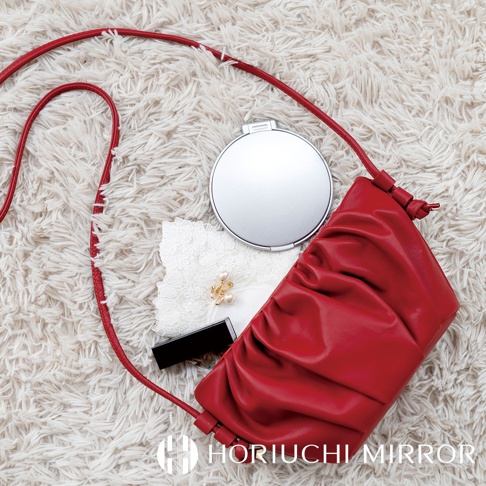 堀內鏡 HORIUCHI MIRROR 的紅色褶皺手拿包，旁邊有一個銀色化妝鏡、化妝品和蕾絲配件，散落在柔軟的地毯上。