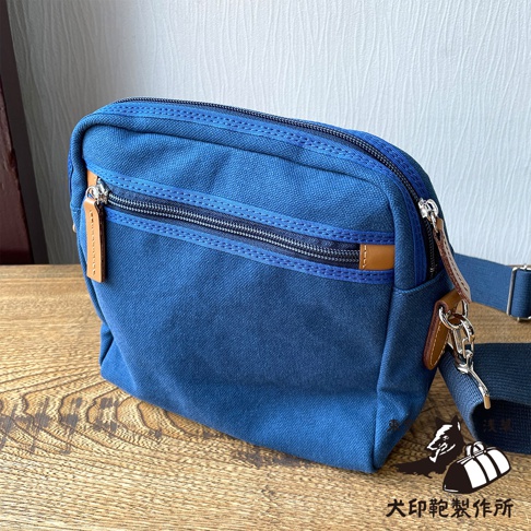 石洗帆布散步包，深藍色，有兩個拉鍊口袋，附有皮質拉環和可調節的肩帶，擺放在木質桌面上。