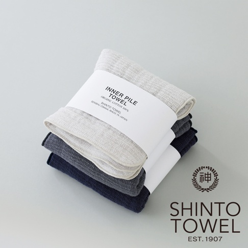三條疊放的 INNER PILE 雙面紗布毛巾，顏色分別為淺灰、深灰和海軍藍。頂部毛巾以白色紙帶包裝，上面印有產品資訊。
