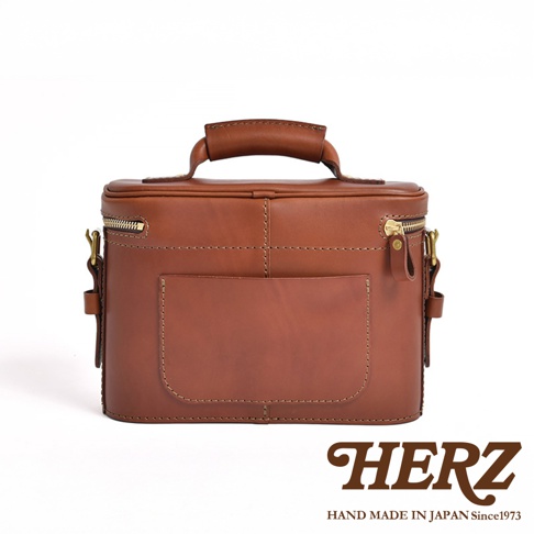 日本HERZ 職人手作皮革厚實相機包 N-8，焦糖色牛皮材質，帶有提把和拉鍊開口，側面有可調節揹帶的 D 型環，正面有一個方形翻蓋口袋，底部有四個金屬腳釘，下方印有 HERZ 品牌標誌與「HAND MADE IN JAPAN Since1973」字樣。