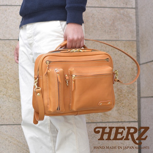 一個棕褐色皮革製成的兩用多功能收納單肩包，包身有多個拉鍊口袋和鈕扣，金色五金配件，附有手提把和可拆卸肩帶。