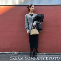 一位女士手持一個米色半月形迷你手提包，包包採用光滑皮革材質，配有頂部提把。她身穿灰色針織外套、黑色洋裝和黑色綁帶靴。