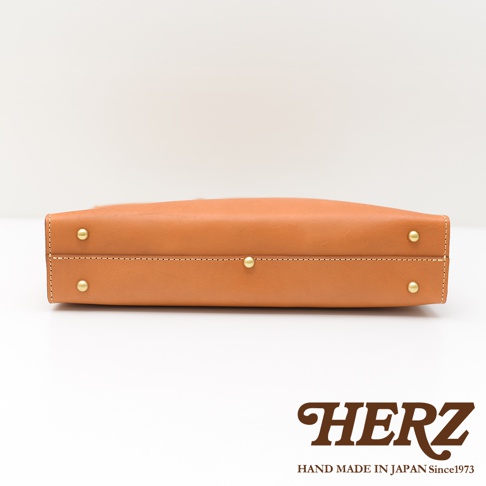 HERZ 品牌標誌，下方為 HAND MADE IN JAPAN Since1973 字樣，中間為棕色皮革公事包的側面，帶有金屬铆釘和縫線細節