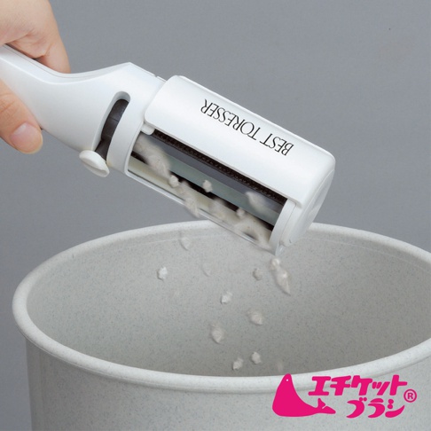 白色免耗材抗菌衣物除塵刷，前端的滾輪上沾有灰白色棉絮，正被傾斜置於一個淺灰色的圓形容器上方，容器內散落著少量棉絮。