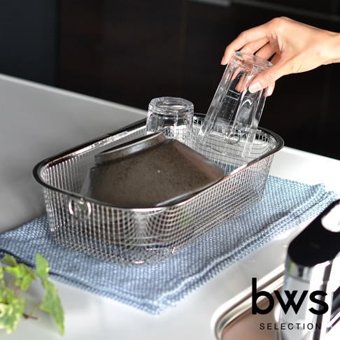 簡約風格的長方形不鏽鋼瀝水籃，內有兩個透明玻璃杯和一個深色碗，瀝水籃置於藍色紋理的布墊上，一只手正將一個玻璃杯放入籃中。
