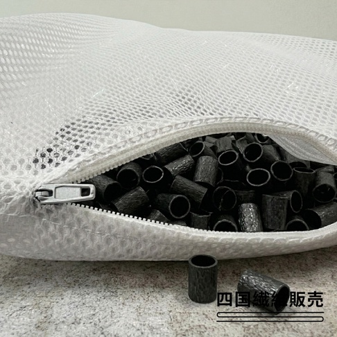 一個裝滿黑色備長炭軟管的白色網狀透氣枕頭，部分軟管散落在枕頭前方，下方標註四國纖維販售字樣。
