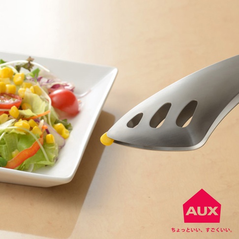 金屬沙拉夾夾著一顆玉米粒，夾身為銀色，有橢圓形鏤空設計，一旁是裝著鮮豔沙拉的白色方形盤。