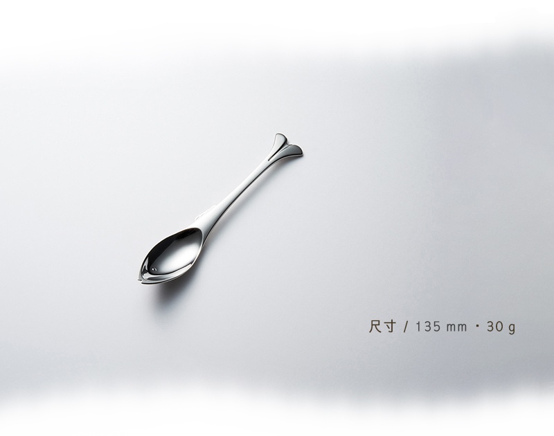 一隻不鏽鋼點心匙，造型獨特，宛如一條小魚，銀色金屬光澤，置於淺灰背景上，右下方有尺寸和重量標示。