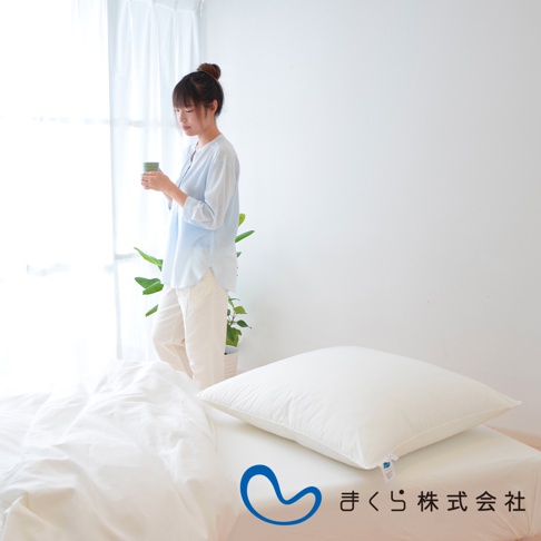 寬大蓬鬆的白色方枕，枕套材質柔軟，放在純白色床單上，旁邊有綠葉植物，一位女士手持杯子站在窗邊。