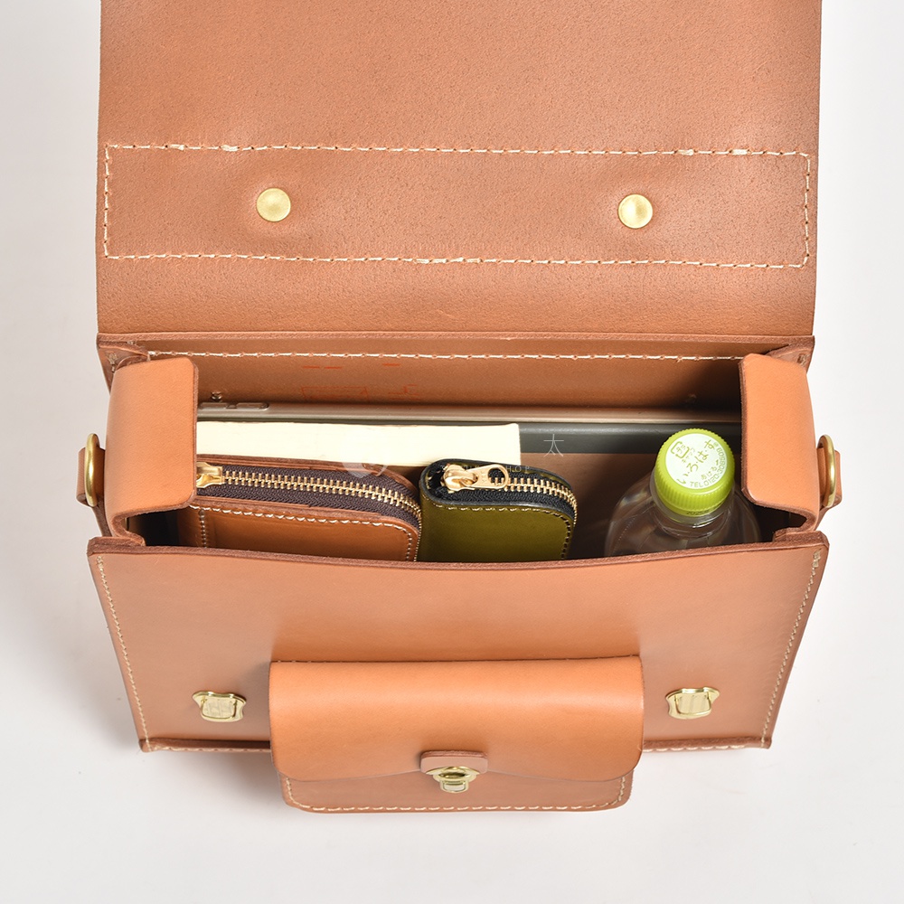 職人手作皮革學院風三用迷你後背包，展示打開的包內空間，可見一本平放的書籍、兩個拉鍊小包（一個棕色，一個橄欖綠）以及一瓶綠色瓶蓋的飲用水。包身為淺棕色皮革，縫線清晰。