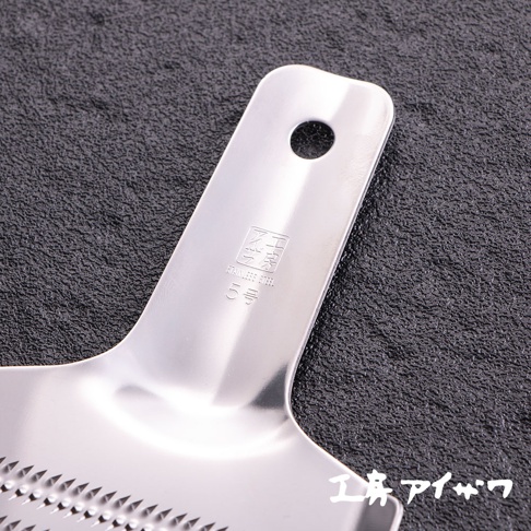 銀色不鏽鋼食材磨泥器，帶有掛孔的長形手柄，手柄上壓印有品牌標誌和「STAINLESS STEEL 5號」字樣。磨泥器部分是平坦的，佈滿鋒利的齒狀磨孔。