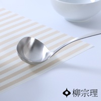 一個中型不鏽鋼湯杓，具有拋光的銀色外觀，放在米白色與淺棕色條紋的墊子上，背景有白色碗的模糊輪廓。