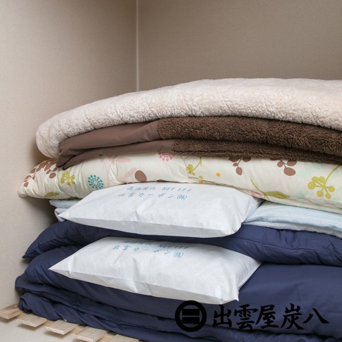 在衣櫃中層層堆疊的棉被與寢具之間，夾放著兩個白色的出雲屋炭八除溼木炭包，包裝表面印有日文字樣與出雲屋炭八的品牌標誌，呈現居家收納與除溼的實景。