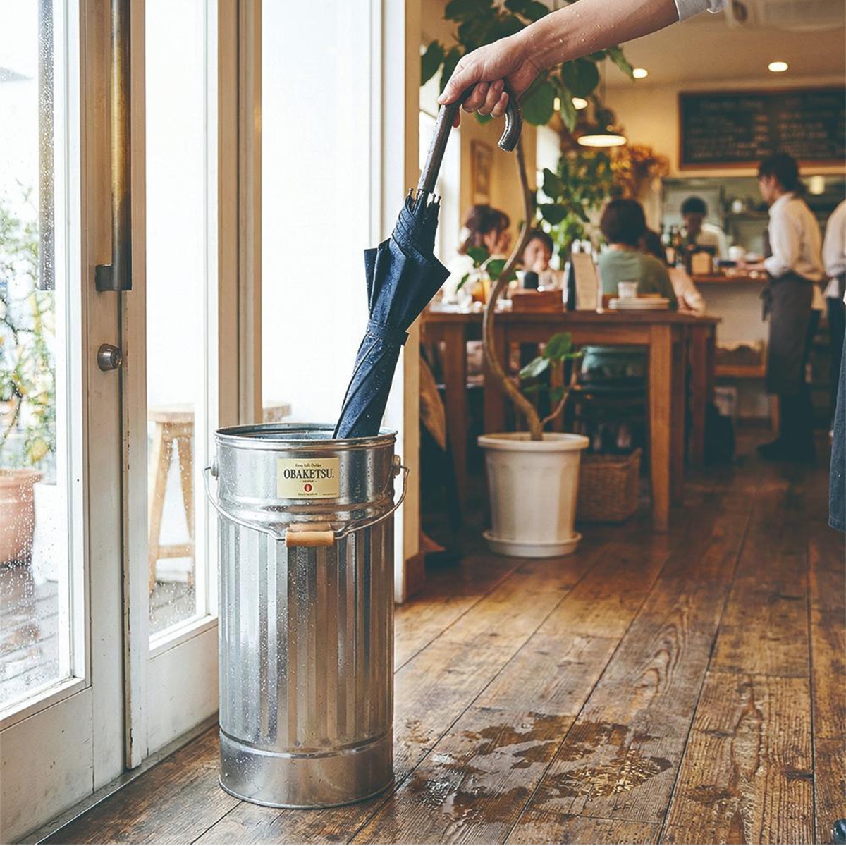 OBAKETSU 手工製鍍鋅鋼傘桶，內部放置深藍色摺疊傘，置於有水漬的木質地板旁，門外陽光灑入室內咖啡廳。