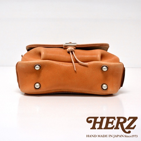 橘色皮革郵差包，有鹿角扣和皮革繩，底部有四個金屬鉚釘。