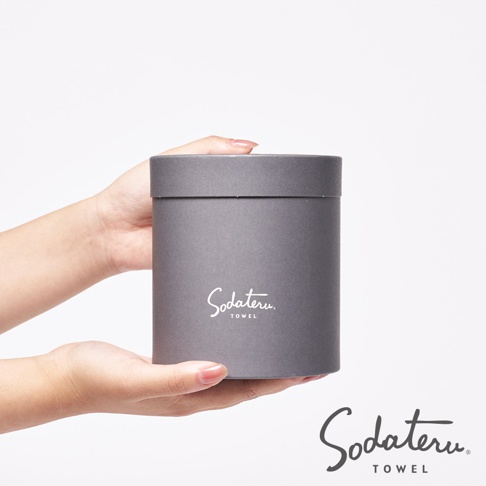 雙手捧著灰色圓筒形禮盒，盒身印有白色「Sodateru TOWEL」字樣。