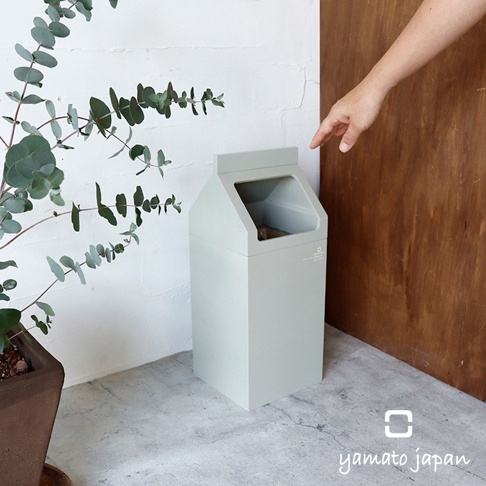 北歐風格淡綠色大開口木製垃圾桶，側面有品牌標誌，置於白色紋理牆壁旁，左側有綠色植物，右側有木質背景。
