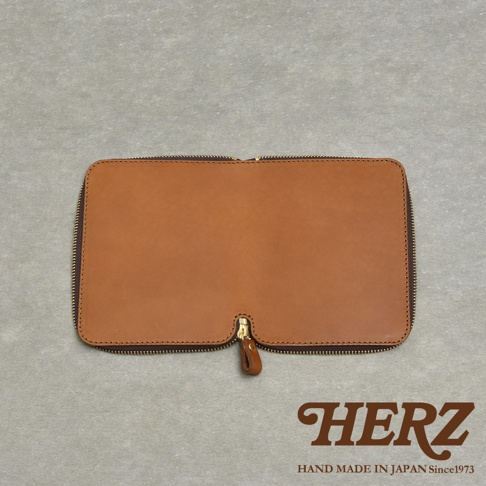 HERZ 品牌標誌，下方有 HAND MADE IN JAPAN Since1973 字樣，中間是一件棕色皮革製的拉鍊式收納包，置於灰色紋理背景上。