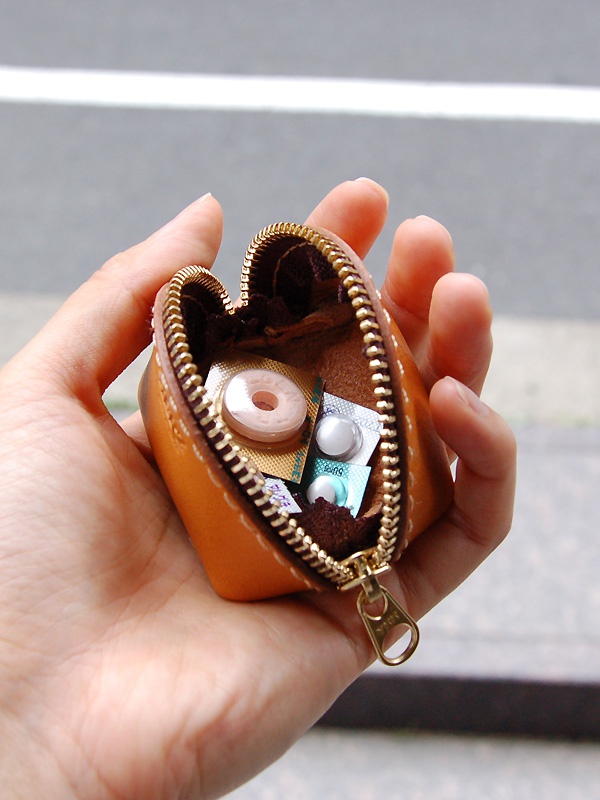 職人手作皮革貝殼造型零錢包，採用橘棕色皮革製成，搭配金色拉鍊，內含數包藥丸與一顆糖果。
