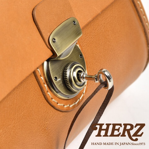 職人手作皮革迷你手提包，特寫其復古黃銅色旋轉鎖扣和棕色皮革材質，細膩的縫線與皮革紋理清晰可見，搭配一條細長的深棕色皮革繫帶，整體展現精緻工藝。