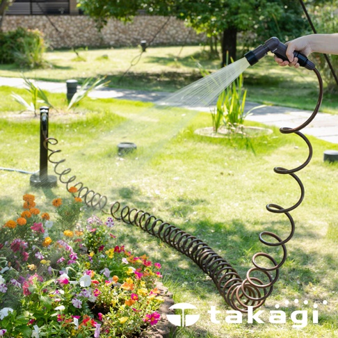 棕色螺旋狀的室外澆花水管，一端由手持，噴灑細緻的水流，另一端連接到草地上的花壇。