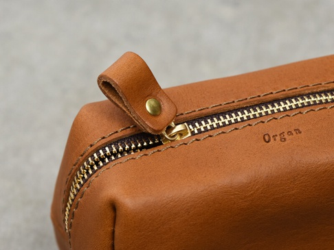 淺棕色皮革製成的職人手作Organ拉鍊收納包，特寫展示了金色拉鍊、皮革拉環和壓印的「Organ」品牌標誌，縫線清晰可見。