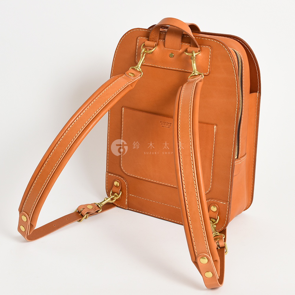 職人手作皮革商務休閒兩用後背包 R-36，展示為淺棕色，採用光滑皮革材質，配有可調節的皮革肩帶、黃銅金屬扣環和拉鍊。包款背面有一個方形的貼袋，上面印有品牌標誌。