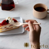 一隻手拿著一支淺色木製點心叉，叉子有三齒，靠近一塊裝飾有鮮奶油、草莓和藍莓的蛋糕。背景是淺色桌面，一旁放著一個木質咖啡杯和一個玻璃茶壺。