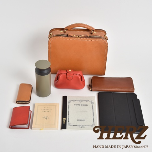 職人手作皮革兩用方形單肩包，搭配一個棕色長夾、一個紅色零錢包、一個綠色保溫瓶、一個棕色卡夾、一個紅色卡夾、兩本筆記本和一個黑色平板電腦保護套。
