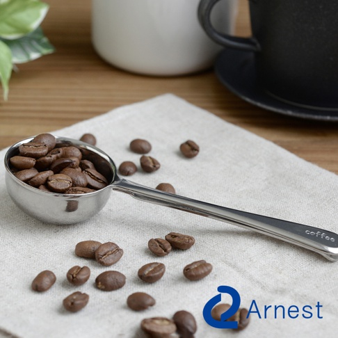 銀色金屬咖啡量匙，帶有夾子功能，量匙內盛有烘焙過的咖啡豆，周圍散落著咖啡豆，量匙把手上印有「coffee」字樣。