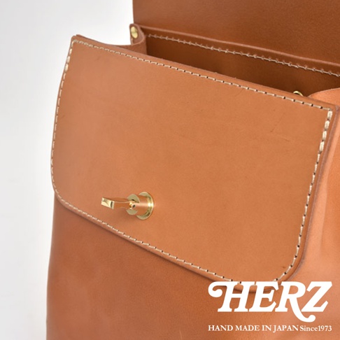 職人手作皮革翻蓋迷你後背包，採用光滑的棕色皮革製成，配有金色旋轉鎖扣，邊緣以白色縫線裝飾。