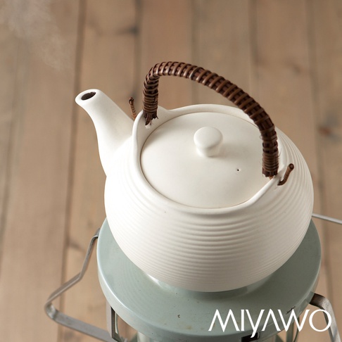白色直火系列陶土茶壺，容量 2L，壺身帶有橫向紋路，配有編織藤柄，壺蓋中央有圓形鈕扣，壺嘴處冒出蒸氣。