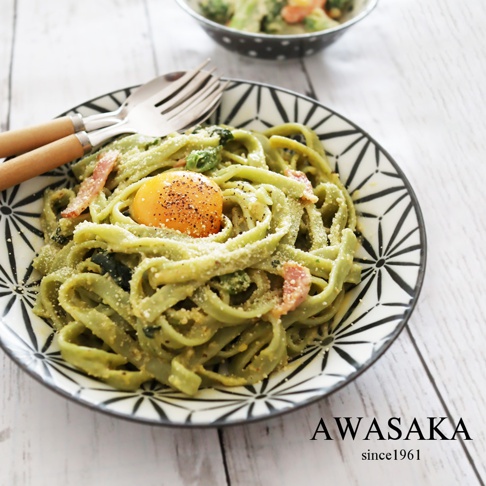 一盤綠色寬麵條，上面放著一顆生蛋黃、培根丁和帕瑪森起司粉，搭配木質手柄的銀色餐具，置於帶有黑色幾何圖案的淺湯盤中，背景為白色木紋桌面。