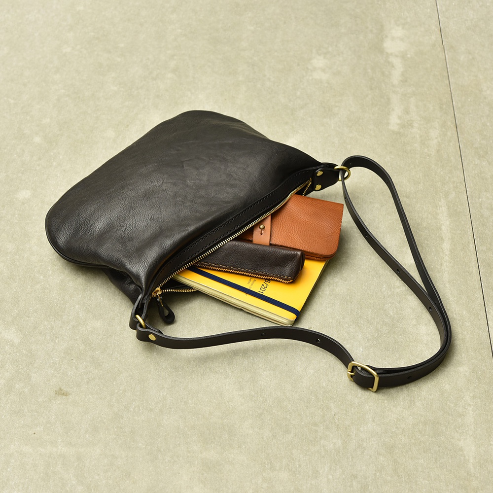 黑色皮革單肩半月包，包身敞開，露出內部的黃色筆記本、咖啡色皮夾和一個深棕色皮製小物。包的肩帶為黑色皮革，配有金色金屬調節環。