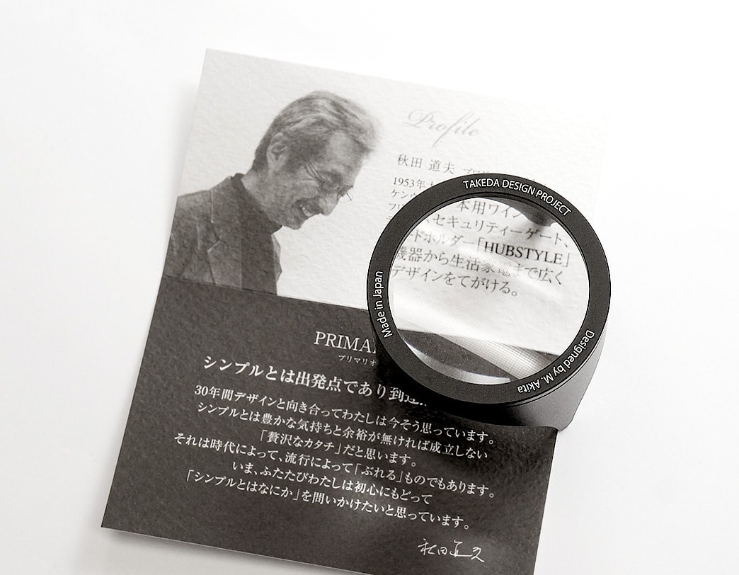 黑色鋁製閱讀放大鏡，鏡環上刻有「TAKEDA DESIGN PROJECT」、「Made in Japan」、「Designed by M. Akita」等字樣，放大鏡置於印有男性肖像及日文文字的卡片上。
