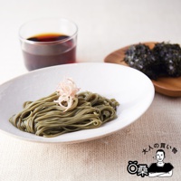 白盤中盛有綠色蕎麥麵，頂部飾有粉紅色薑絲，旁邊擺著一杯褐色液體和裝有兩團黑海苔的木製小碟。