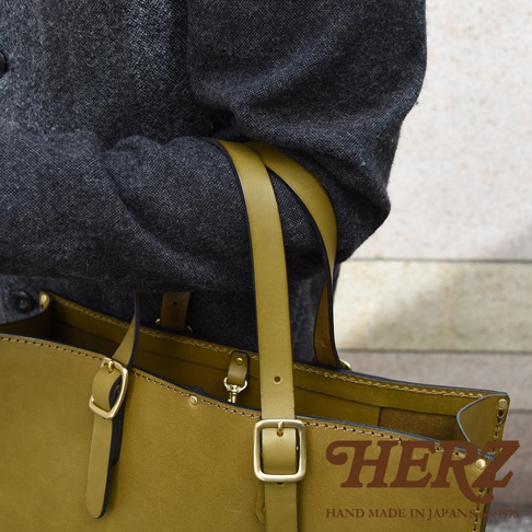 一件橄欖綠色皮革托特包，配有同色系皮革提把及金屬扣環，由職人手工縫製，呈現簡約商務風格。