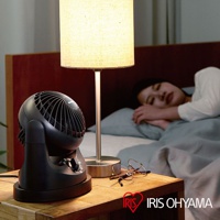 黑色迷你型空氣循環扇，帶有可調節的旋鈕，放置在床頭櫃上，旁邊有一盞檯燈和一副眼鏡，背景是睡覺的女性。